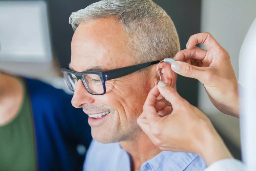 woman's hands placing behind the ear hearing aid on senior man's ear