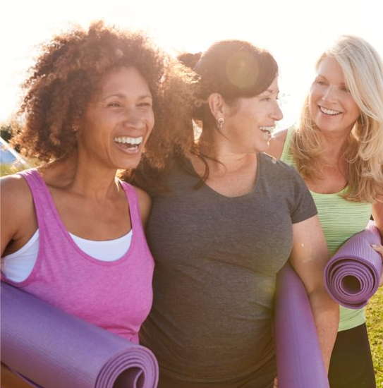 Three laughing women carrying yoga mats in the sunlight