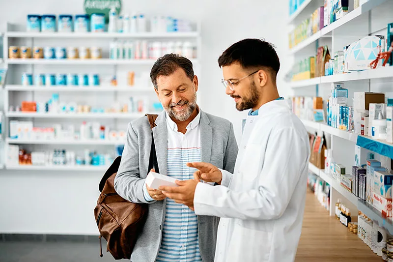 Bearded man with shoulder bag looks at box with man in white coat at a pharmacy. The pharmacist is pointing to the product while customer smiles. 