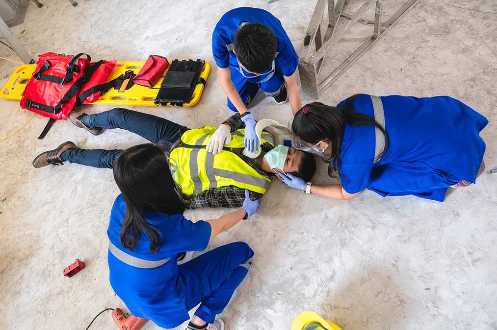 Three people in blue smocks and pants giving first aid to injured, masked worker on his back. A stretcher and a ladder on its side are nearby.