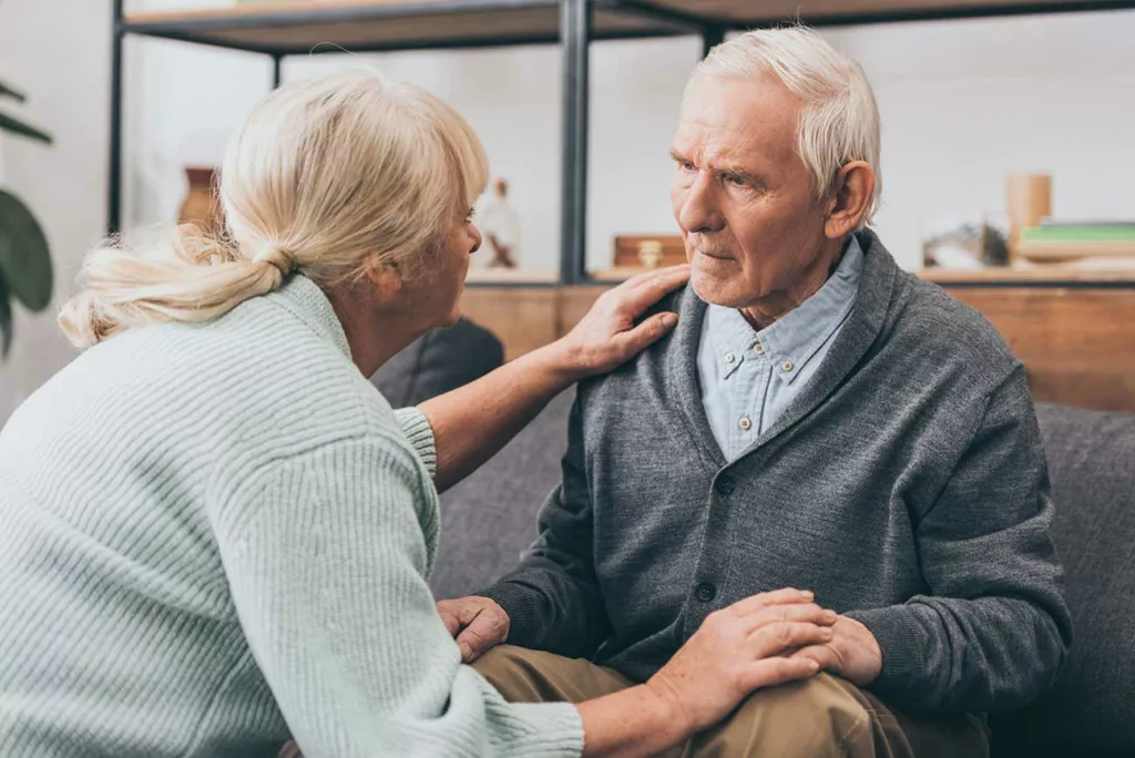 Senior couple. She's kneeling in front of him with hands on his. He's sitting on couch with hands on knees, looking confusedly at her. 