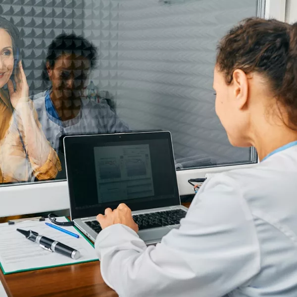 middle-aged woman holding button attached to cable in audiology sound booth, smiling through window at tech, who has hands poised on laptop.