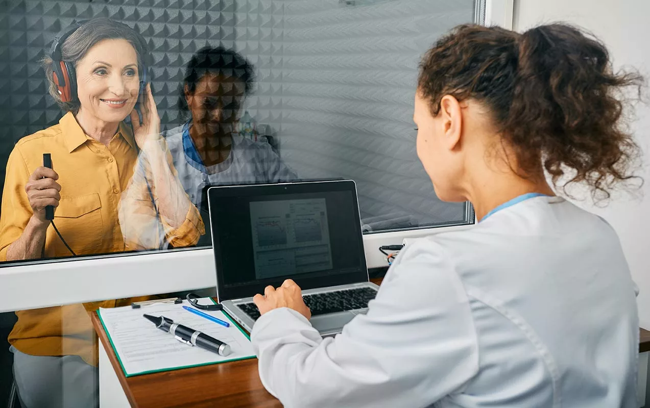 middle-aged woman holding button attached to cable in audiology sound booth, smiling through window at tech, who has hands poised on laptop.