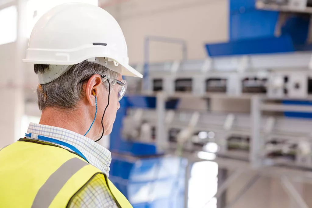 Man in his 50s wearing white hard hat, yellow safety vest looking at bank of machines while also wearing glasses and earplugs. 