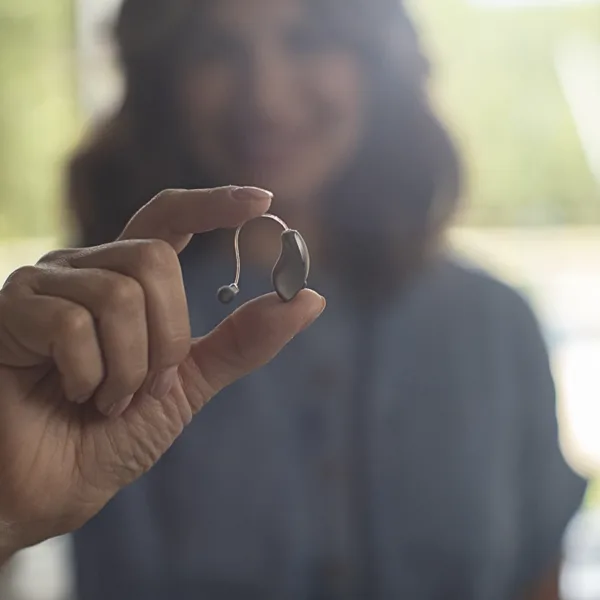 A women holding up a small behind-the-ear BTE hearing aid, her face is slightly blurry in the background