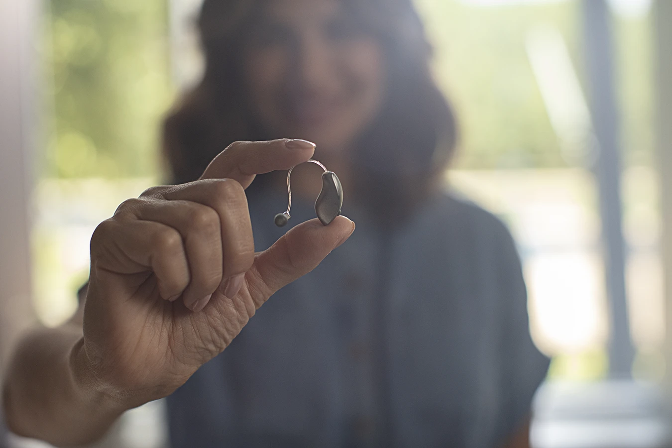A women holding up a small behind-the-ear BTE hearing aid, her face is slightly blurry in the background
