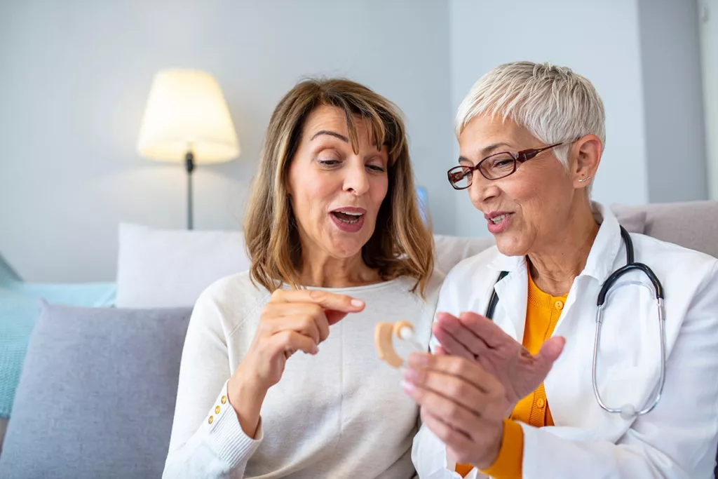 woman in lab coat holding hearing aid, sitting next to senior woman who is pointing at it and speaking animatedly