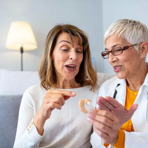 woman in lab coat holding hearing aid, sitting next to senior woman who is pointing at it and speaking animatedly