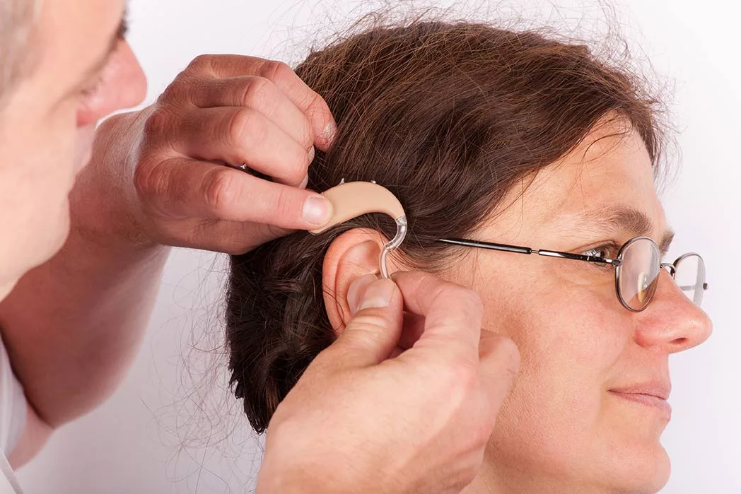 Man's face in profile, and his hands, as he fits hearing aid on woman. She's wearing glasses and has her dark red hair in a bun. 