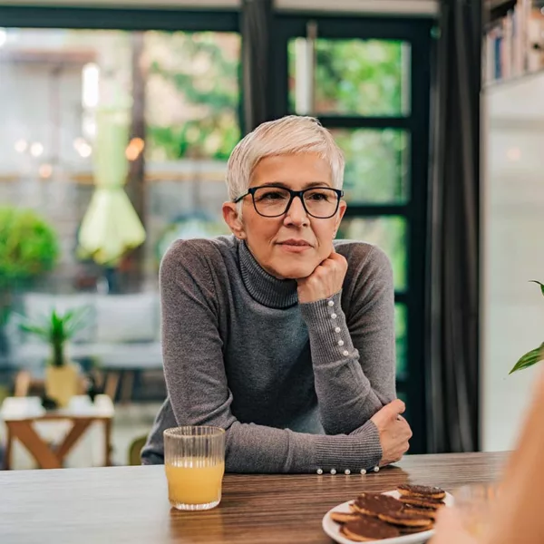 Gray-haired woman with short hair cut, glasses, and turtleneck sweater has her hand on chin, leaning in to listen to friend at table.