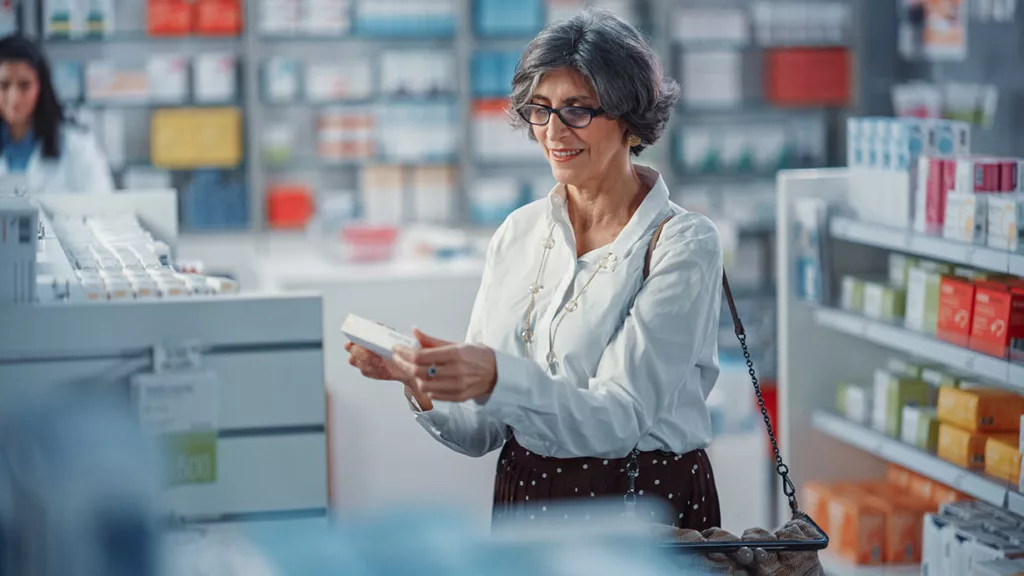 Older woman with glasses holding, and smiling at, small package in a drug store.