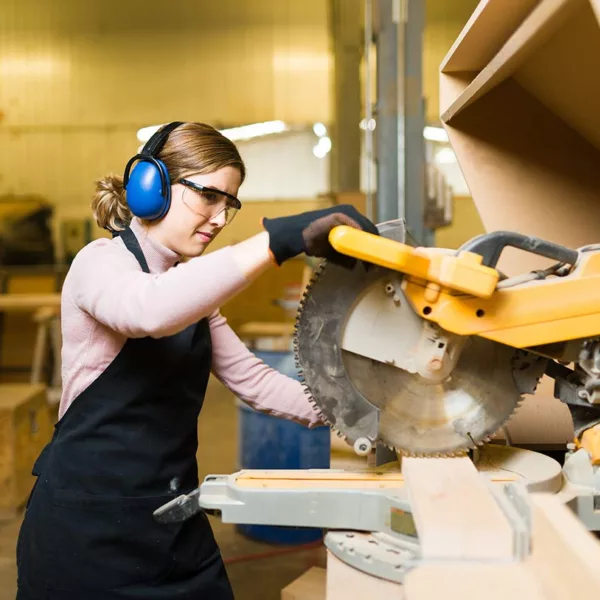 Woman in overalls and long-sleeved shirt wearing ear protection while operating a table saw.
