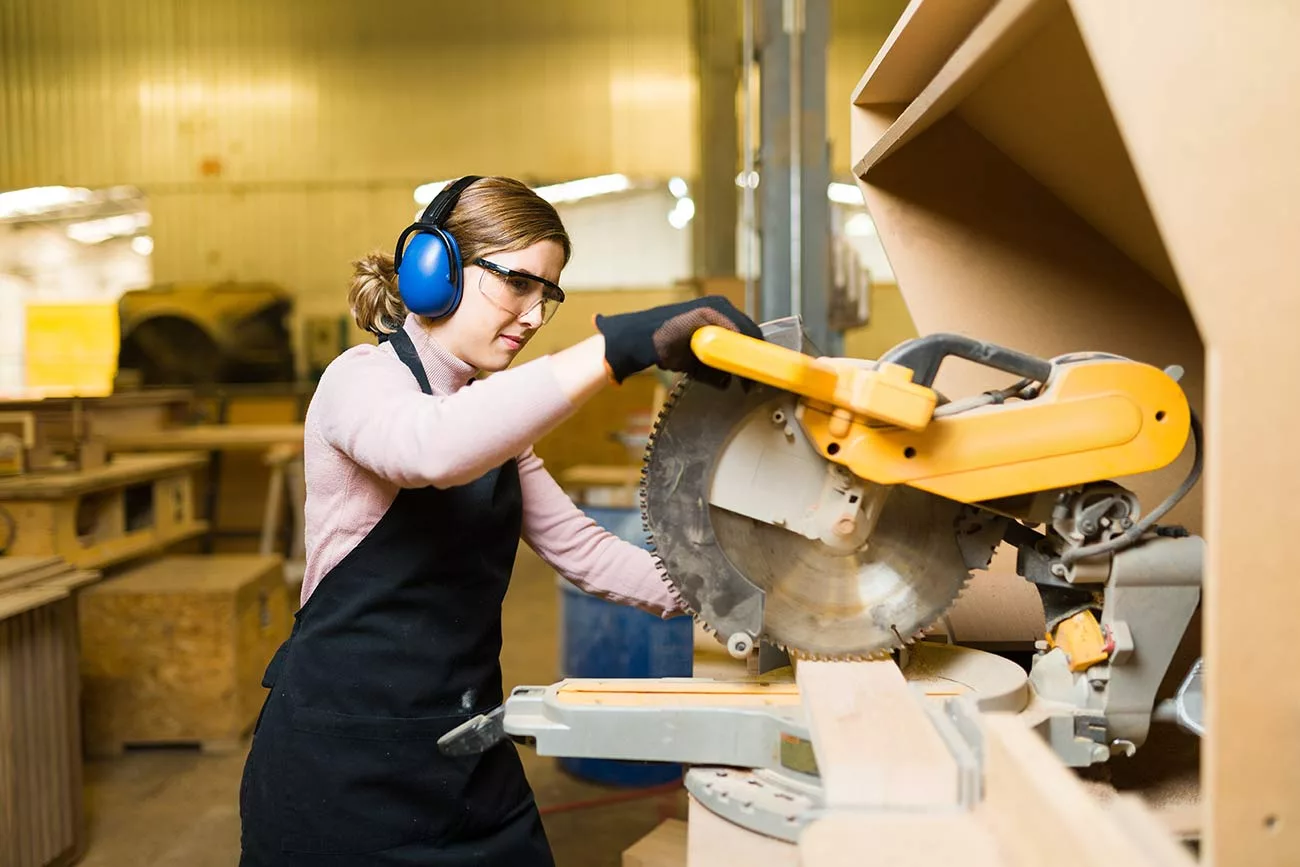 Woman in overalls and long-sleeved shirt wearing ear protection while operating a table saw.