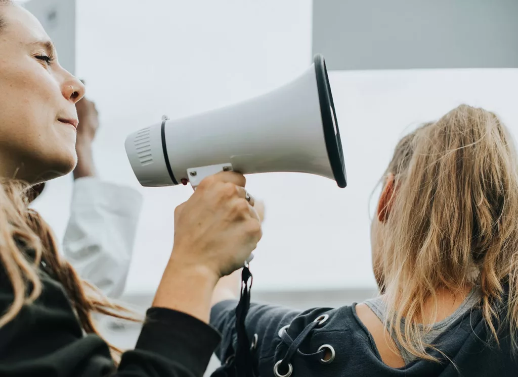 woman speaking into bullhorn at outdoor gathering with another woman near the bell