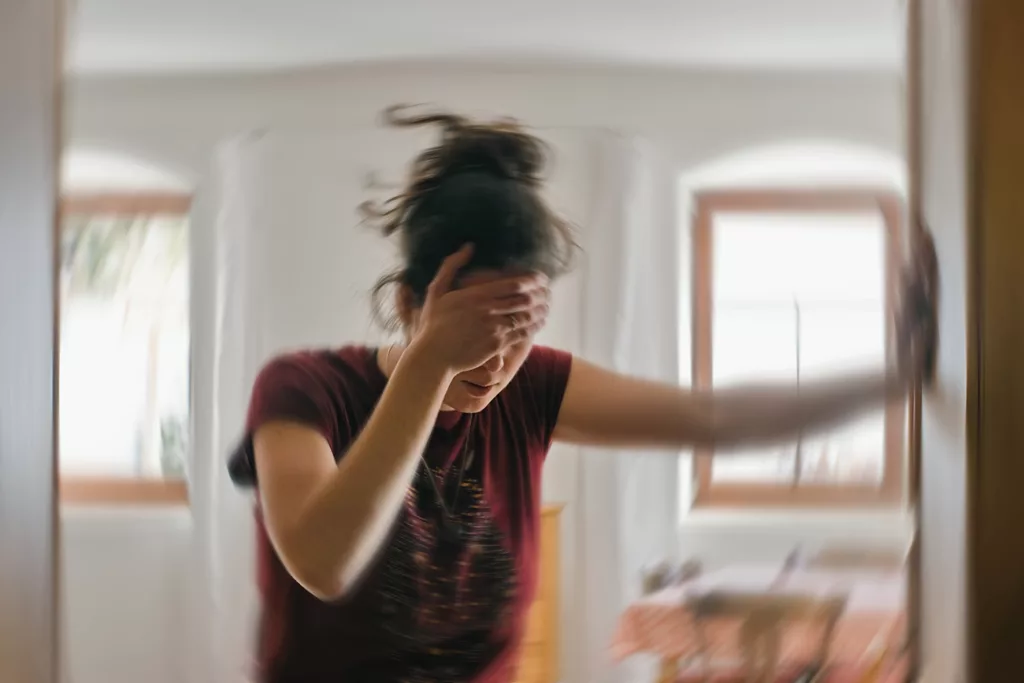 woman standing, bracing with hand on wall, and holding her head with other hand. Blurred to indicate dizziness.