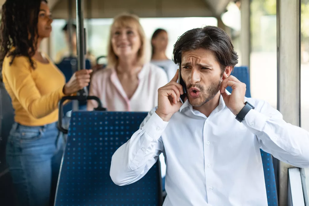Man looking confused and seeming to say "who" while holding phone to one ear and holding other ear while on bus