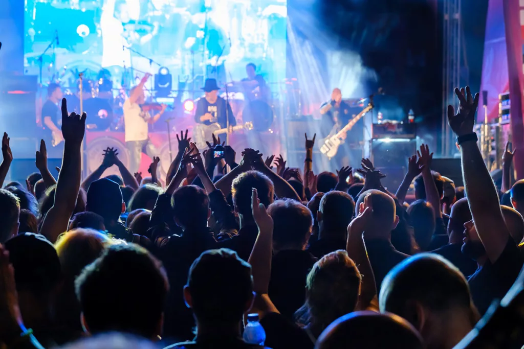 A crowd of concert goers clapping with their hands in the air and singing along with a large stage with a rock band out of focus in the background 