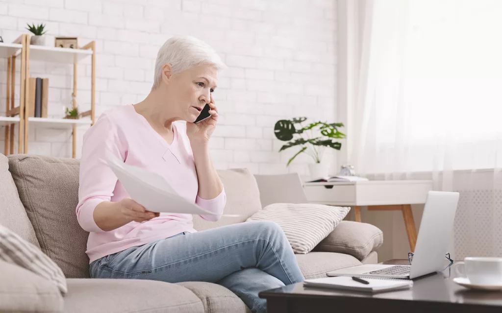 Confused senior on couch holding papers in one hand, and mobile to ear with other, looking at laptop on coffee table