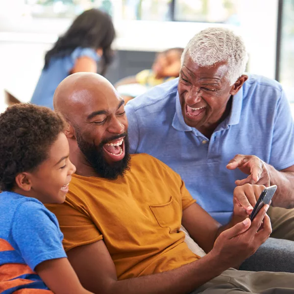 Grandfather wearing hearing aids, father, and grandson laughing and looking at cell phone while sitting on sofa