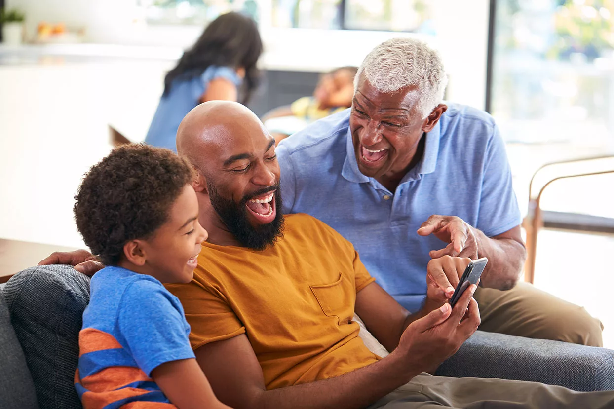 Grandfather wearing hearing aids, father, and grandson laughing and looking at cell phone while sitting on sofa