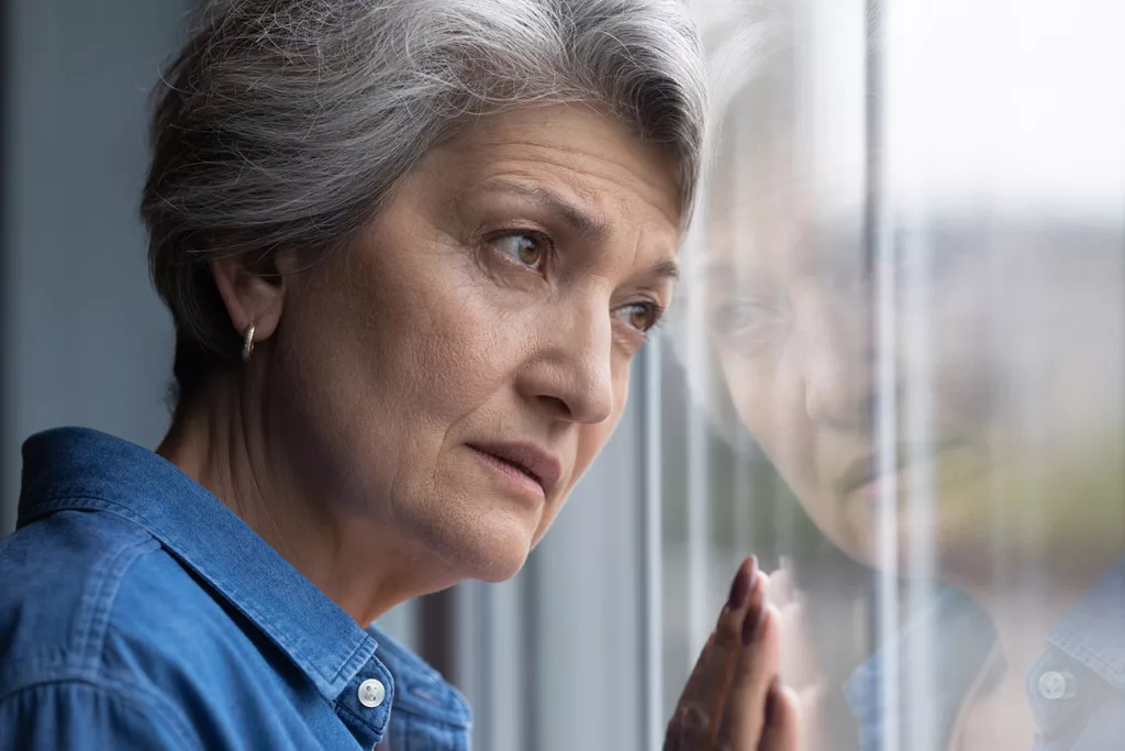 Senior woman looking through window with hand to glass and sad expression. 