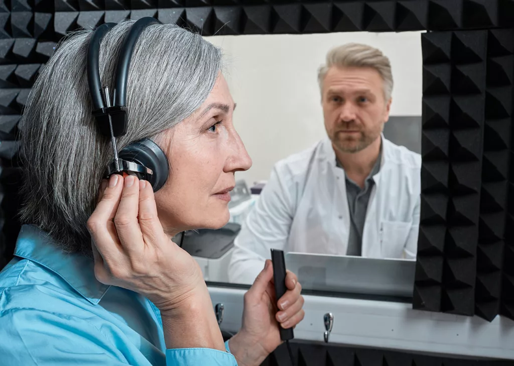 Woman with headphones on in sound booth with clicker. Audiologist seen through window.