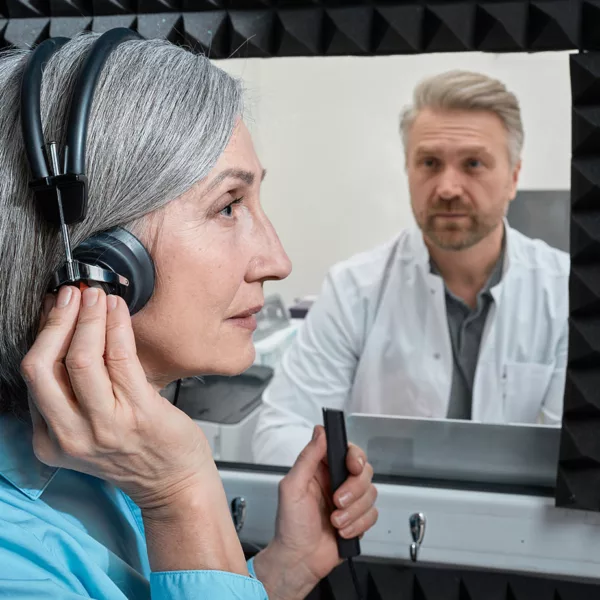Woman with headphones on in sound booth with clicker. Audiologist seen through window.