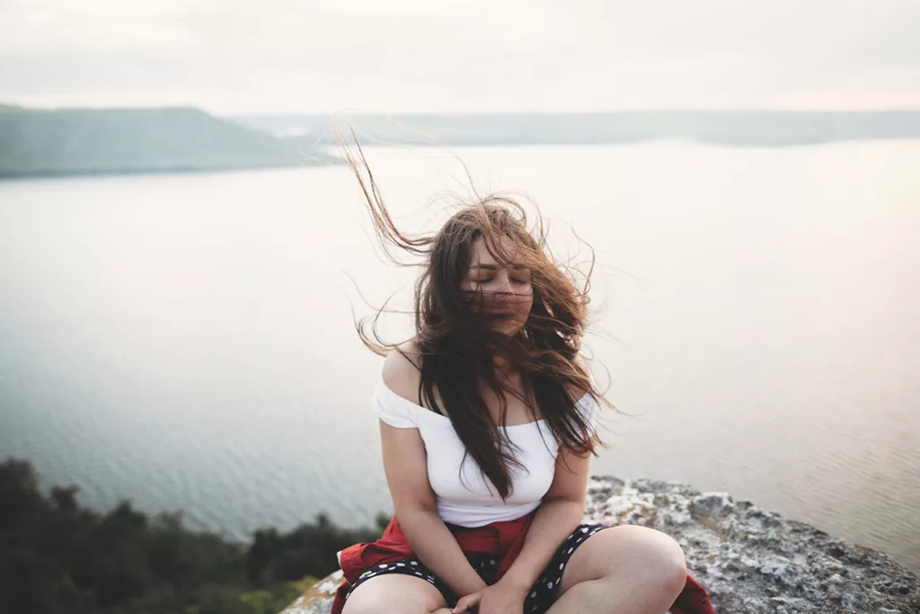 Young woman with windblown hair sitting on outcropping of rock with her back to the ocean and her eyes closed. 