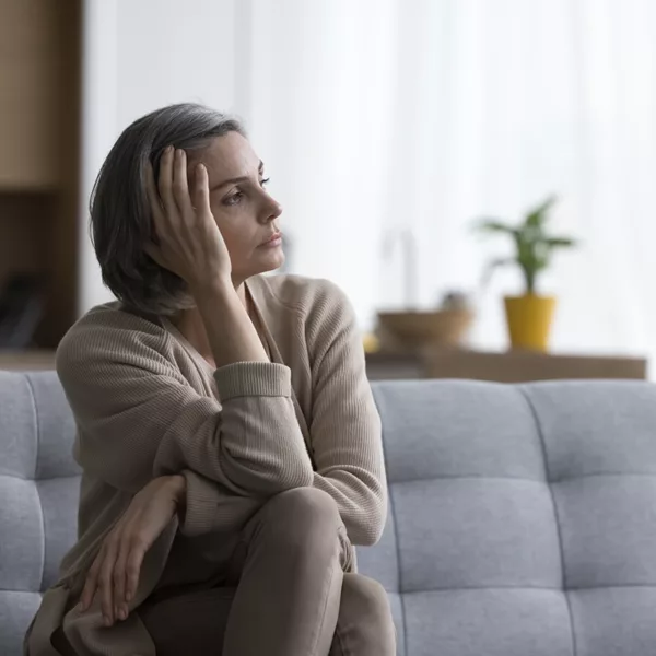 Middle aged short haired woman sitting on sofa at home, looking at window away in deep unhappy thoughts, leaning head on hand, touching face, suffering from depression caused by untreated hearing loss