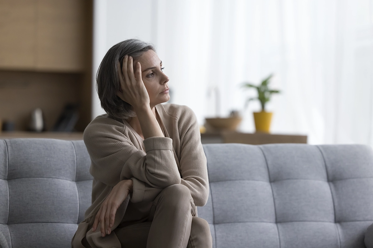 Middle aged short haired woman sitting on sofa at home, looking at window away in deep unhappy thoughts, leaning head on hand, touching face, suffering from depression caused by untreated hearing loss