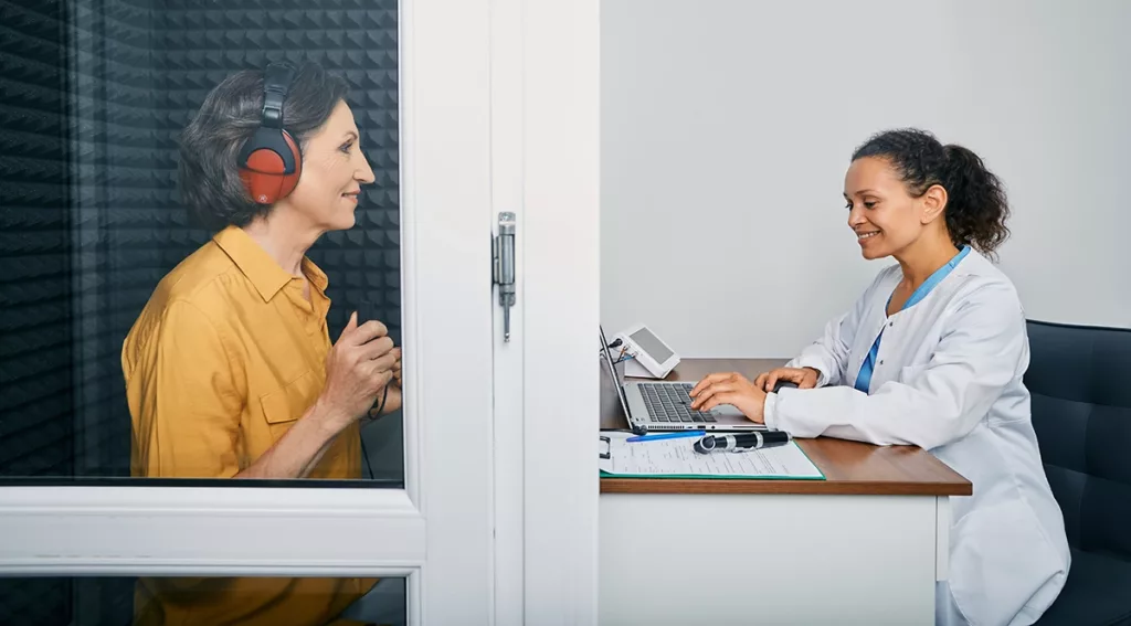 An adult women in a soundproof booth taking a hearing test conducted by a hearing specialist who is sitting at a desk outside the booth smiling