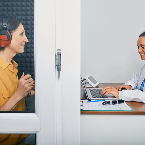 An adult women in a soundproof booth taking a hearing test conducted by a hearing specialist who is sitting at a desk outside the booth smiling
