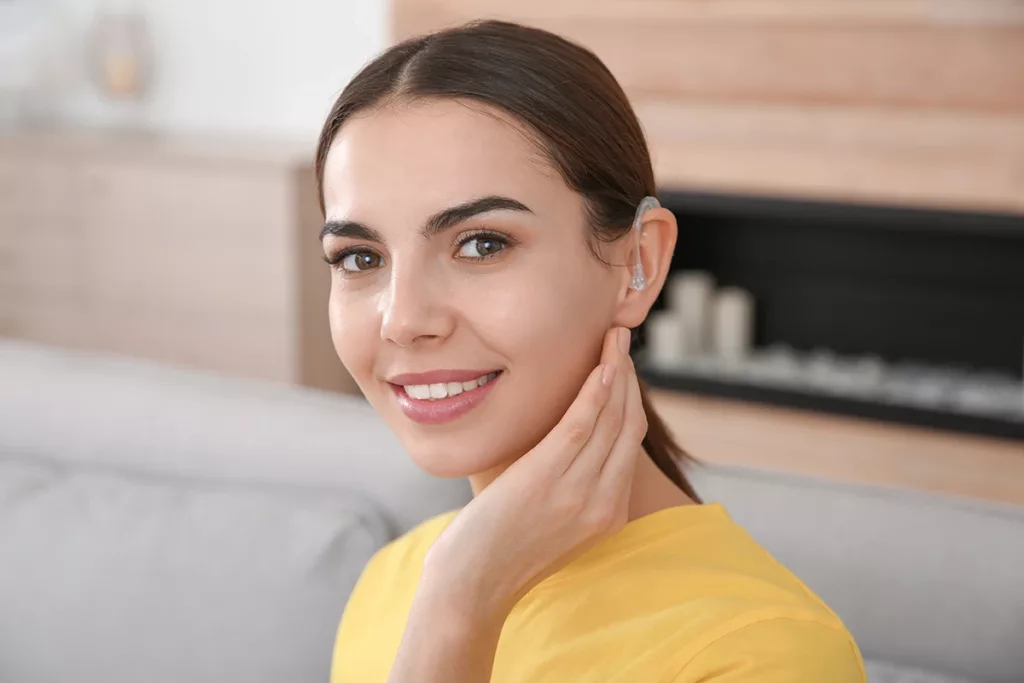 A young woman with brunette hair in a low ponytail, wearing a behind-the-ear (BTE) hearing aid and smiling 