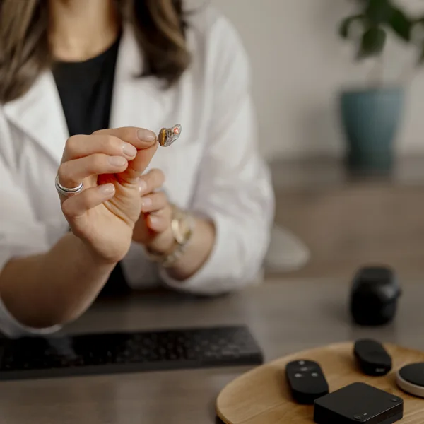 A hearing care specialist holding up a small in-the-ear ITE Bluetooth hearing aid next to a selection of various wireless hearing aid accessories