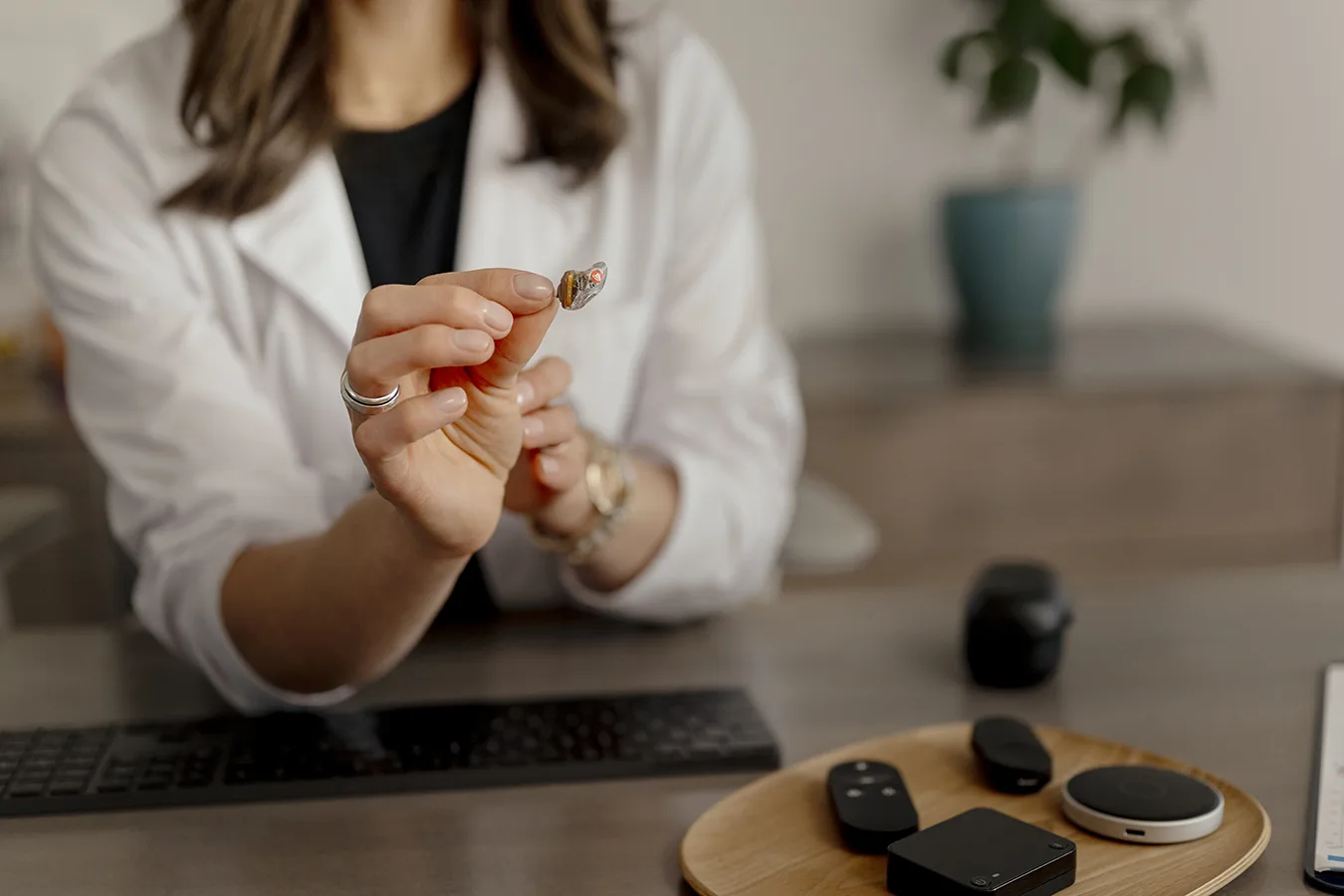 A hearing care specialist holding up a small in-the-ear ITE Bluetooth hearing aid next to a selection of various wireless hearing aid accessories