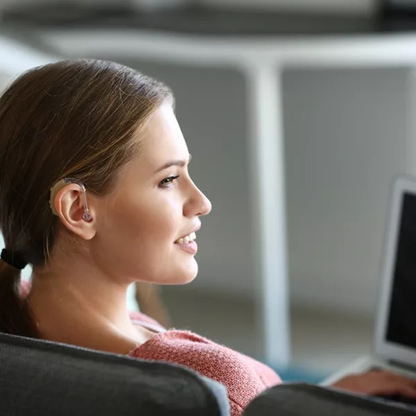 A young women with hearing loss sitting in her living room using wireless hearing aid accessories to stream audio directly from her laptop to her hearing aids.