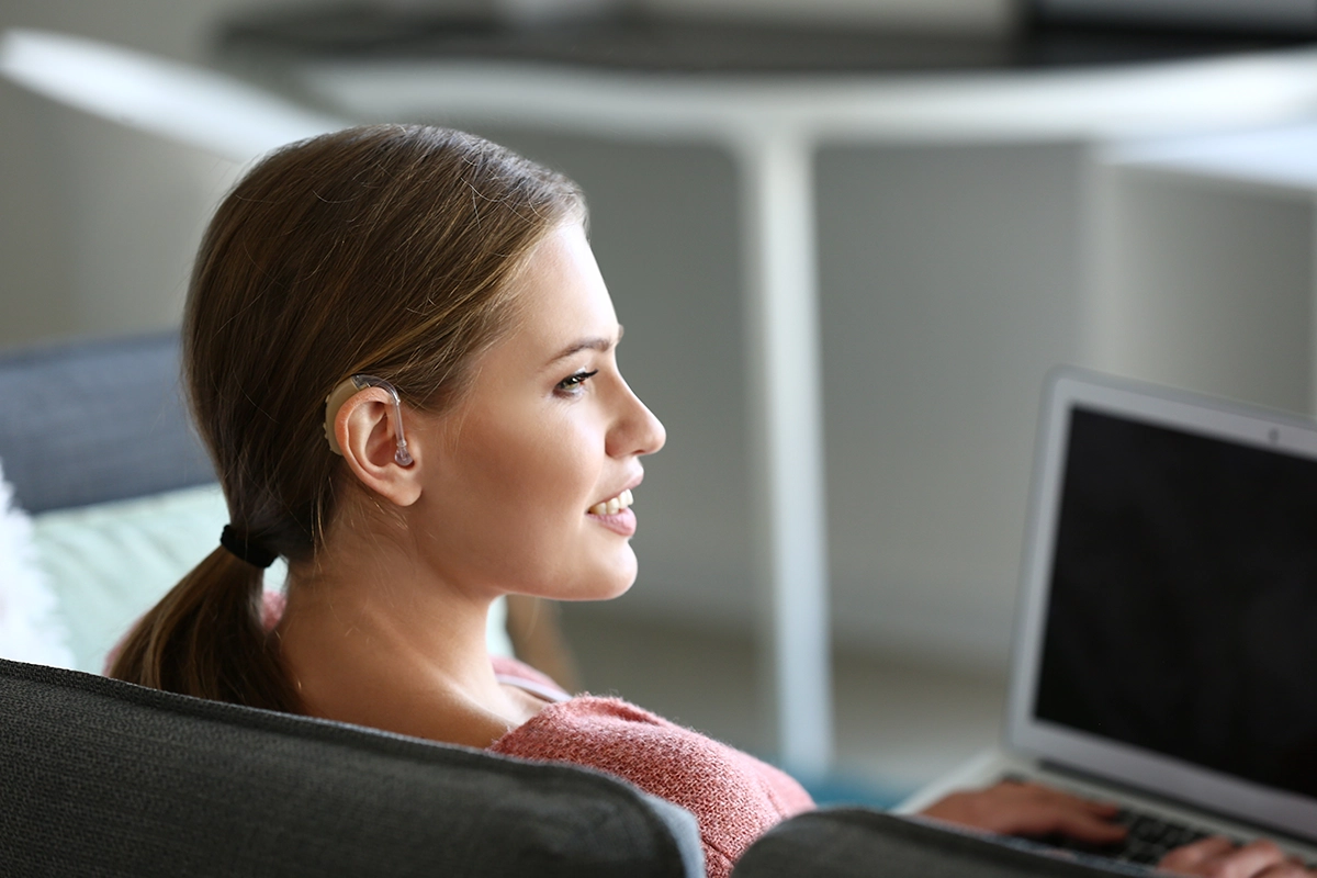 A young women with hearing loss sitting in her living room using wireless hearing aid accessories to stream audio directly from her laptop to her hearing aids.