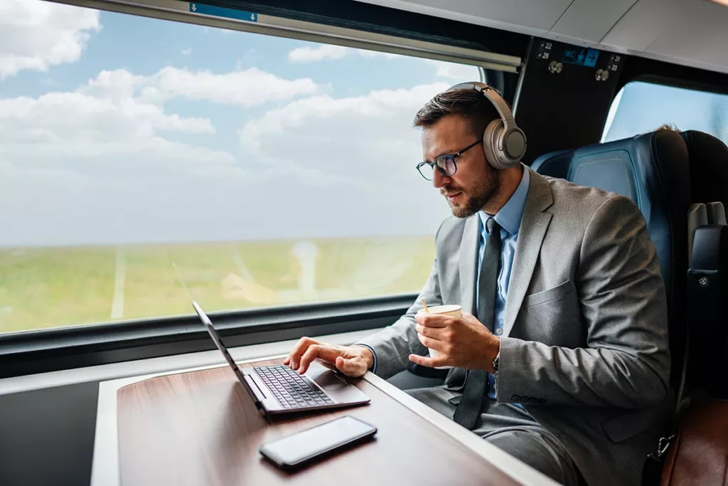 Middle age businessman using his laptop computer with wireless noise-canceling headphones while commuting to work on a train.
