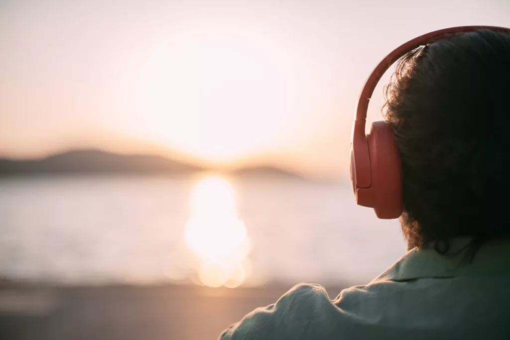 A person sitting on the beach wearing wireless noise-canceling headphones looking out at sunset over the water.