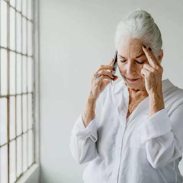 A senior woman with untreated hearing loss talking on the phone by a window in a white room
