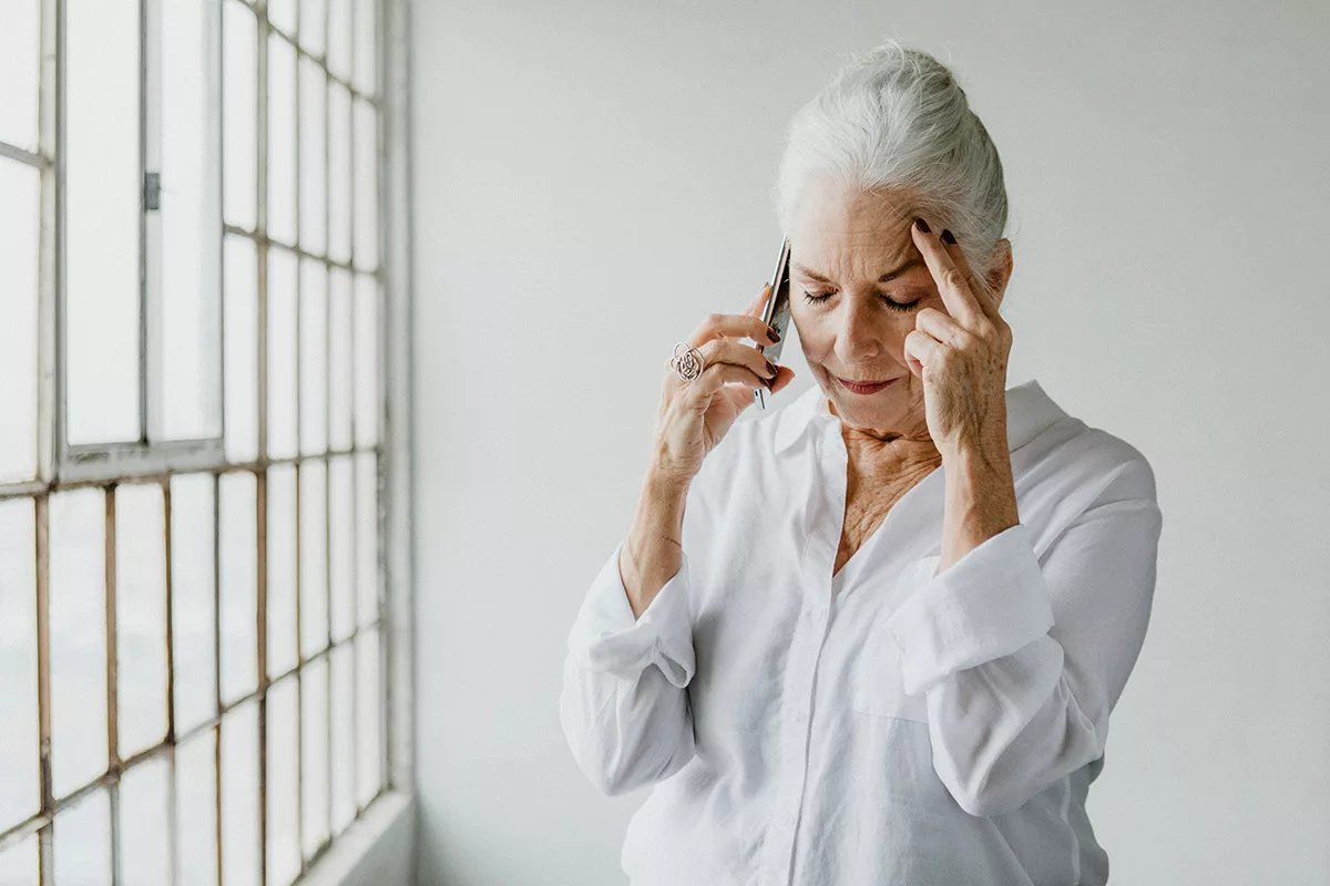 A senior woman with untreated hearing loss talking on the phone by a window in a white room