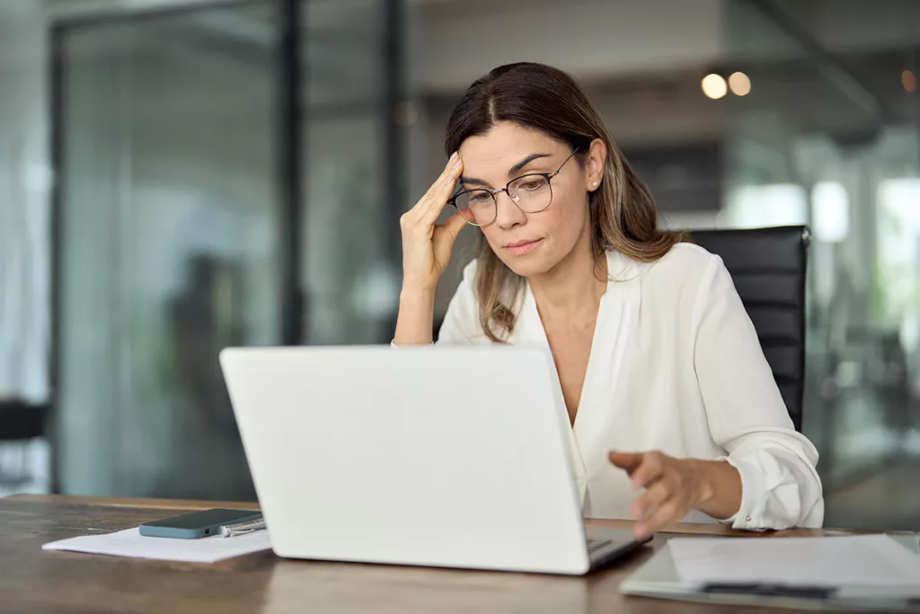 A business woman with untreated hearing loss at work, sitting in front of a white laptop in a business office.