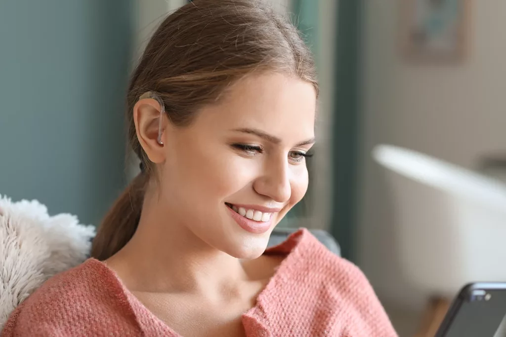 A young women sitting in her living room smiling while she uses her Bluetooth hearing aids to to listen to audio from her smartphone