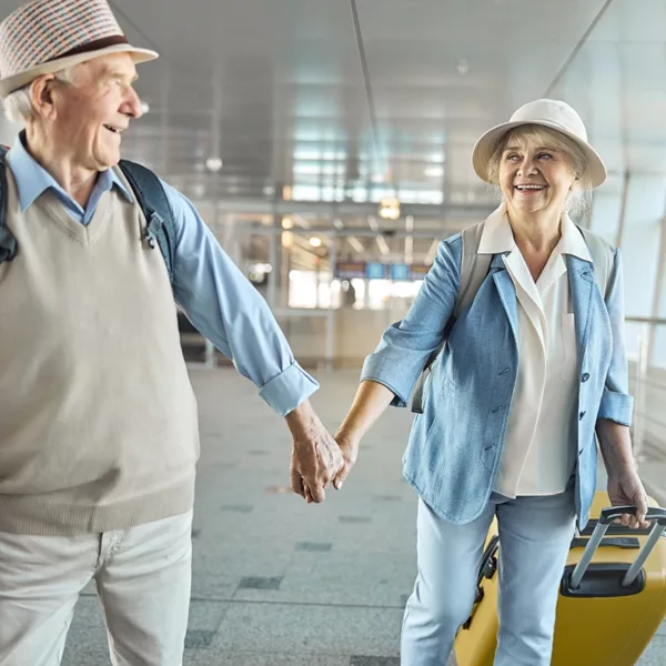 Senior couple happily holding hands and smiling at each other as they walk through an airport with luggage.