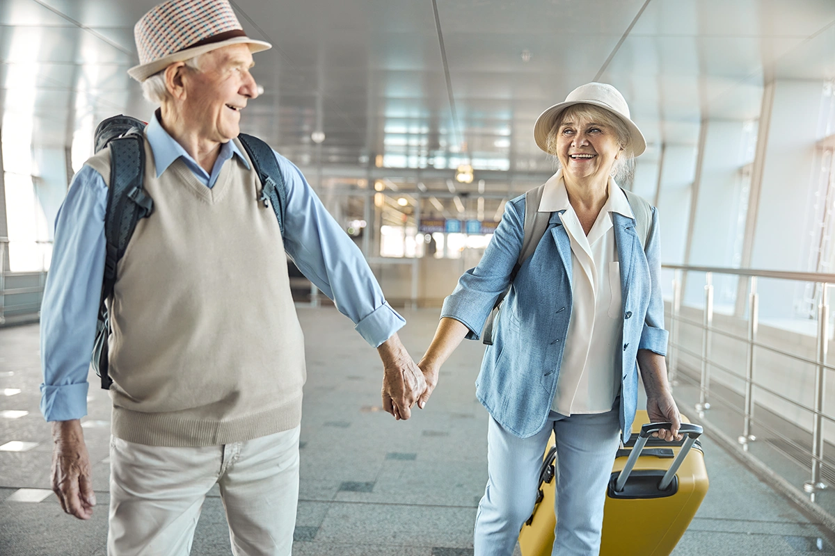 Senior couple happily holding hands and smiling at each other as they walk through an airport with luggage.