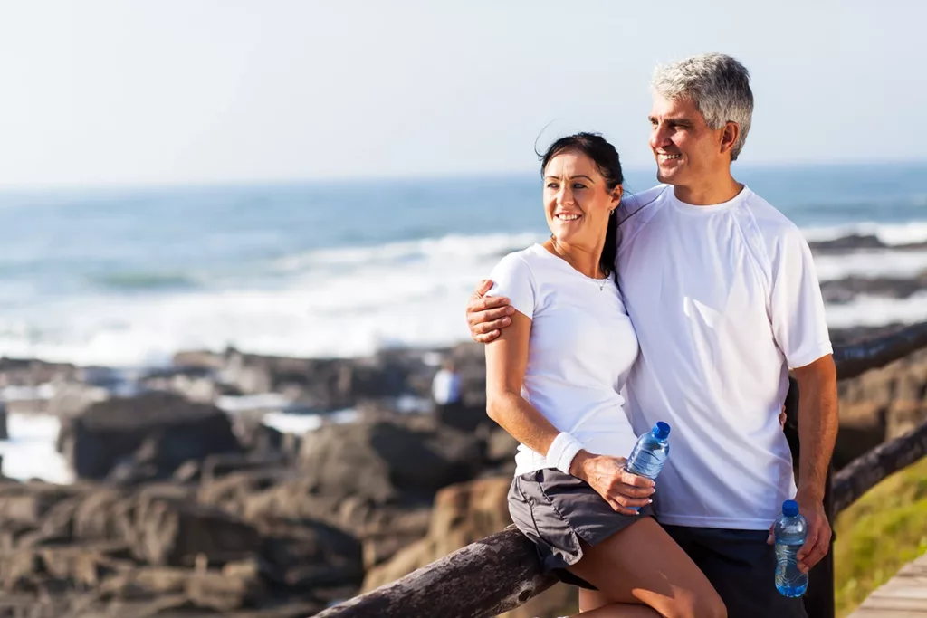 A healthy and happy middle aged couple dressed in athletic, workout clothing, looking out at a shoreline while on a hike together near the beach