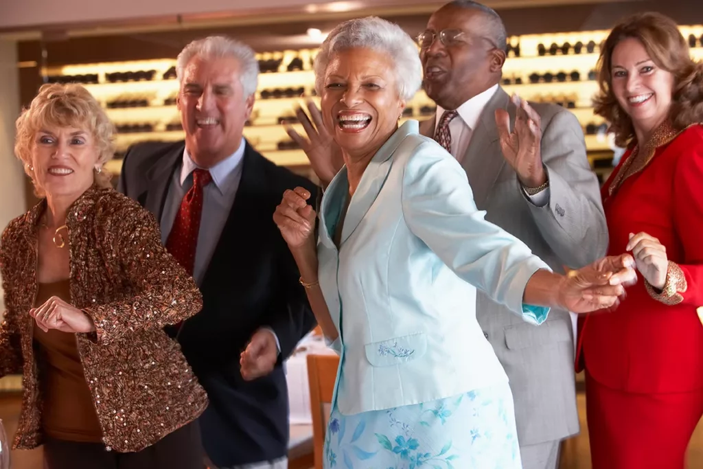 A friend group of older adults, dressed in formal clothes dancing and laughing at a party 