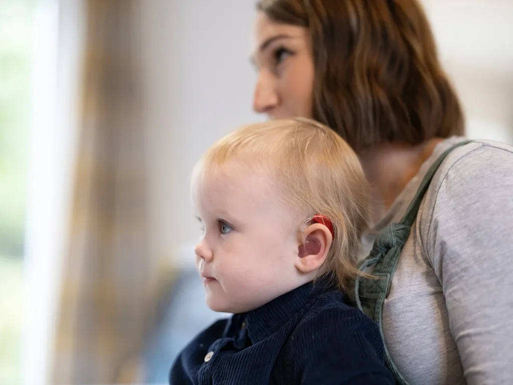 A toddler with non-syndromic hereditary hearing loss wearing a hearing aid, sitting on their mother's lap in a hearing health clinic.