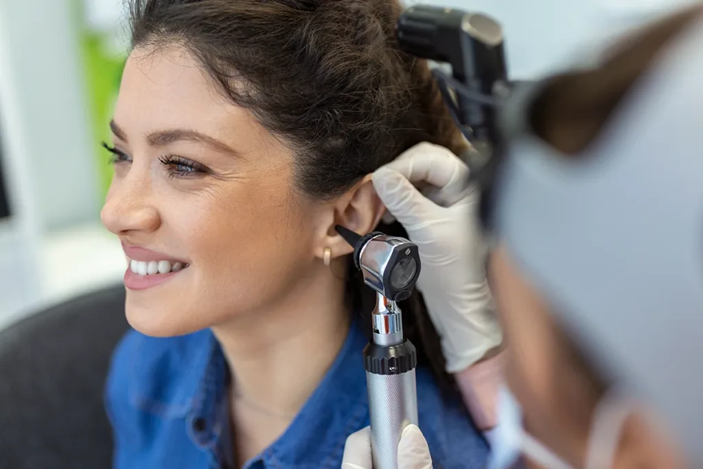 A middle aged women happily sitting in a hearing clinic, while a hearing health care specialists exams her ears with an otoscope 