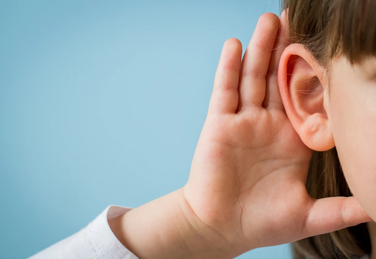 A close up of a small child holding their hand up to their ear as if to help them hear better, symbolizing hereditary hearing loss.